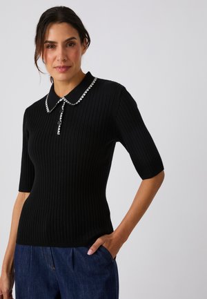 Mujer con una camiseta negra ajustada de manga corta con canalé y cuello negro con ribete blanco, combinada con vaqueros azules, de pie con una mano en el bolsillo.