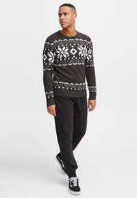 Dark brown knitted sweater with white winter-themed patterns, round neckline, ribbed cuffs, worn with black sweatpants and black sneakers.