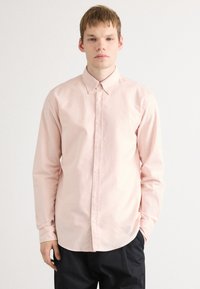 Ung mann står mot hvit bakgrunn i en lys rosa skjorte med knapper og svarte bukser, med den ene hånden i lommen.
