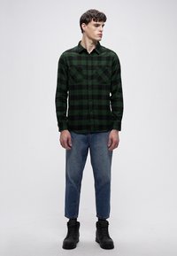 Groen-zwart geruit flanellen overhemd met een knoopsluiting aan de voorkant, borstzakken en een losse pasvorm, gecombineerd met taps toelopende blauwe jeans en zwarte laarzen.