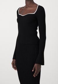 Kvinna klädd i en åtsittande svart ribbad långärmad topp med en vitkantad fyrkantig halsringning och en matchande ribbad kjol.