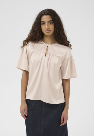 Mladá žena s dlhými kučeravými vlasmi, ktorá má na sebe bledoružovú blúzku s krátkym rukávom a výstrihom na uzol a tmavú rifľovú sukňu.