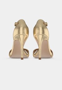 Kaks shiny-kultaa korkeakorkoiset sandaalit nilkkanyörillä, nähtynä takaa yksinkertaisella valkoisella taustalla.