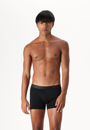 Schwarze Herren-Boxershorts aus glattem Stoff, mit einem gerippten Bund und einer enganliegenden Passform, gezeigt an einem Modell vor einem weißen Hintergrund.