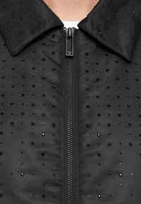 Fekete cipzáras dzseki sima textúrával, amelyen egy nagyméretű gallér található, és elszórt fekete flitterek díszítik.