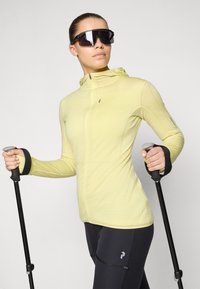 Svetlo rumena zip-up jakna z teksturirano tkanino, prilegajoče oblike in kapuco, v kombinaciji s črnimi športnimi hlačami in pohodniškimi palicami. Nosijo se temna sončna očala.