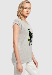T-shirt grigio a maniche corte con una stampa grafica nera e gialla di un personaggio con accessori, abbinato a pantaloni bianchi.