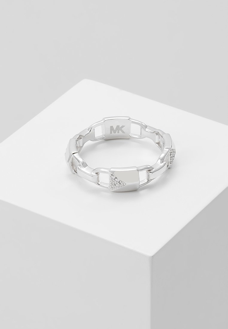 Bague à maillons en argent avec accents diamant triangulaires et logo MK, exposée sur un piédestal géométrique blanc sur fond gris.