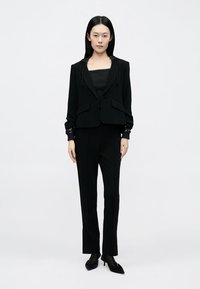 Juoda pritaikyta džemperio striukė su aptakiu siluetu, derinama su ilgomis juodomis kelnėmis. Savybės – minimalistinis dizainas ir struktūruoti pečiai.