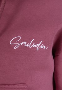 Felpa bordeaux realizzata in morbido tessuto, con il logo "Smilodor" ricamato in bianco. Include una tasca frontale e polsini a costine.