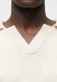 Pull en maille de couleur crème avec un col en V, des bords côtelés et une surface texturée. Le tissu semble doux et léger.