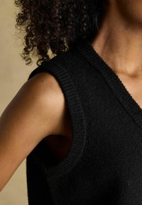 Czarny bezrękawnik dzianinowy z dekoltem w kształcie litery V. Tkanina jest strukturalna z prążkowanymi krawędziami, co nadaje mu ogólnie miękki wygląd.