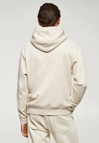 Homme portant un sweat à capuche crème et un pantalon assorti, debout de dos avec une main dans la poche, sur un fond uni.