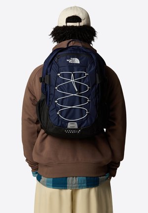 Mørkeblå ryggsekk med sorte detaljer, med elastisk snor-design, justerbare stropper og The North Face-logo på fronten og bunnen.