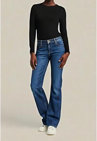 Personne portant un haut noir ajusté à manches longues, un jean bleu taille haute avec une légère évasée, et des chaussures blanches, debout devant un fond uni.