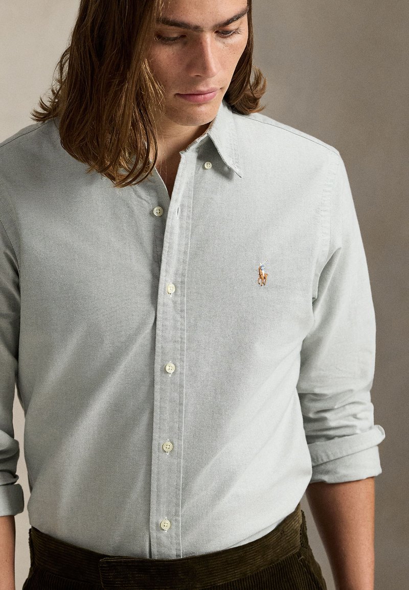 Ανοιχτό γκρι πουκάμισο με κουμπιά, με απαλό υφή και μικρό κεντημένο λογότυπο στο αριστερό στήθος, φορεμένο με τα μανίκια σηκωμένα.