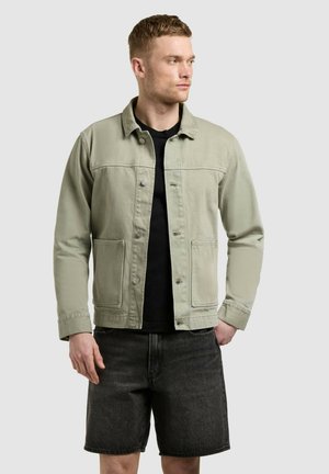 Mann trägt eine hellgrüne, geknöpfte Jacke über einem schwarzen Hemd und schwarzen Jeansshorts, schaut nach rechts vor einem einfarbigen grauen Hintergrund.