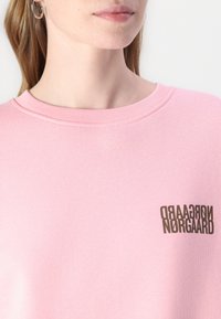 Roza dukserica s okruglim izrezom, s smešnim logotipom od smeđeg teksta u vertikalnom rasporedu na levoj strani. Meko, rebrasto teksturo.