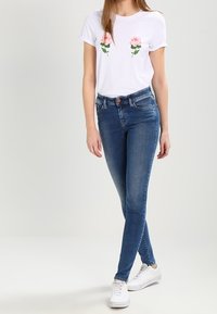 Vit t-shirt med rosa blommor broderade framtill, tillsammans med blå skinny jeans och vita sneakers, allt mot en ljus bakgrund.