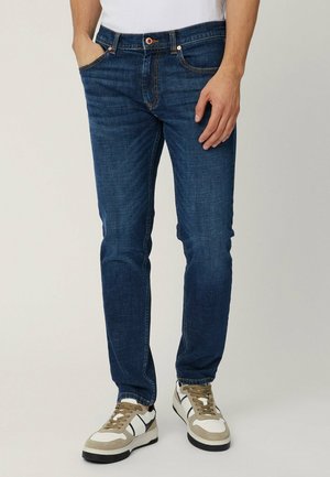 Jeans in denim blu scuro con vestibilità dritta, design a cinque tasche, cuciture arancioni e chiusura a bottone in vita. Indossati con sneakers bianche.