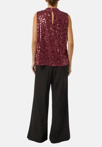 Burgunderfarbenes, ärmelloses Oberteil mit Pailletten, das einen Schlüssel-Ausschnitt auf der Rückseite hat. Kombiniert mit weiten schwarzen Hosen. Glatte Textur und glänzende Akzente.