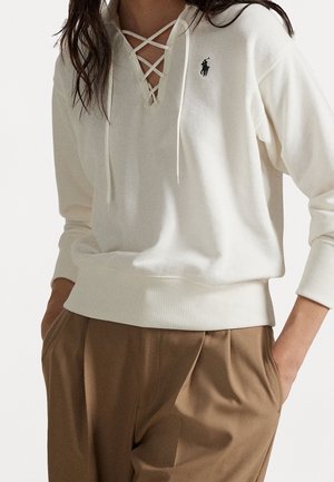 Femme portant un sweat à capuche blanc à lacets avec un petit logo et un pantalon plissé marron, mains dans les poches.