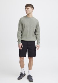 Hellgrüner gerippter Langarmpullover, schwarze Cargo-Shorts mit Seitentaschen, graue Sneakers und weiße Crew-Socken. Einfaches, lässiges Outfit.