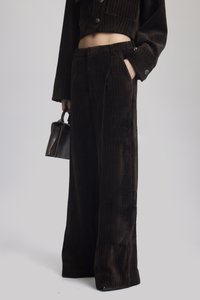 Světlé široké kalhoty z kordu v tmavě hnědé barvě s texturovaným proužkovým vzorem, bočními kapsami a čistým lemem. Stylizované s malou kabelkou.