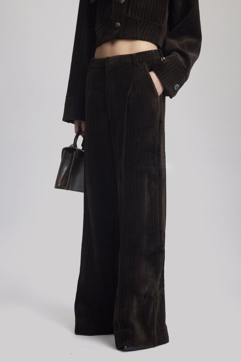 Světlé široké kalhoty z kordu v tmavě hnědé barvě s texturovaným proužkovým vzorem, bočními kapsami a čistým lemem. Stylizované s malou kabelkou.