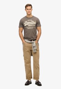 Camiseta gráfica marrón con el texto "SuperDry", chinos beige, suéter gris atado a la cintura, mocasines oscuros, tono de piel claro.