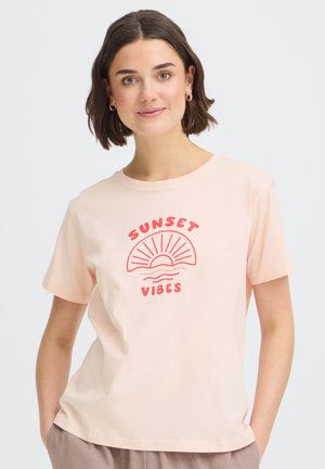 Jeune femme aux cheveux courts et foncés portant un t-shirt rose clair "Sunset Vibes", debout, les mains dans les poches, devant un fond uni.
