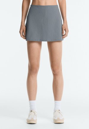 Kvinde iført en sort og hvid mini-nederdel med lodrette striber, hvide sokker og hvide sneakers, stående mod en ensfarvet hvid baggrund.