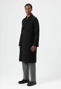 Schwarzer Wollmantel mit einem Reverskragen und langen Ärmeln, versehen mit einem seitlichen Schnallendetail. Kombiniert mit grauen Hosen und schwarzen Schuhen.