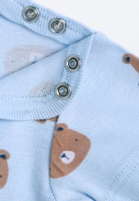 Světle modrý textilní overal s potiskem medvídků a stříbrnými druky na límci. Měkká textura a jemné pletené detaily jsou viditelné.