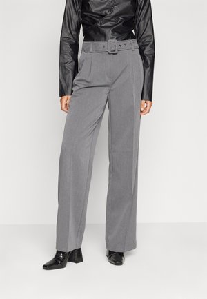 Femme portant un pantalon gris taille haute à jambes larges avec une ceinture assortie, un top en cuir noir à manches longues et des chaussures à talons noires.