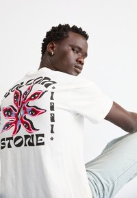 White short-sleeve T-shirt featuring a colorful eye pattern graphic on the back and bold text reading "Volcom Stone." Soft, casual fabric.