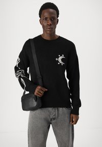 Pulover negru tricotat cu un model alb de lună pe piept și motive albe complicate pe mâneci. Asortat cu blugi gri și o geantă neagră.