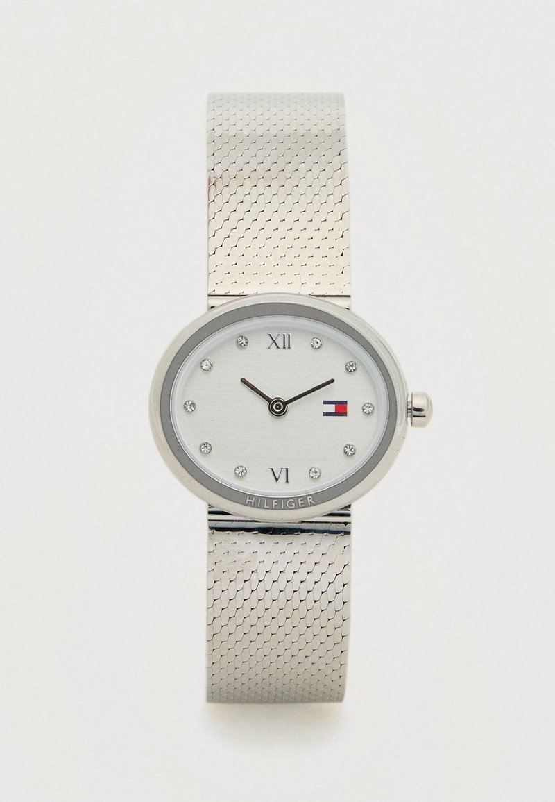 Zilveren polshorloge met een gaasarmband, een ovale wijzerplaat, kristallen uurmarkeringen, Romeinse cijfers op 12 en 6, en een klein vlaglogo op de wijzerplaat.