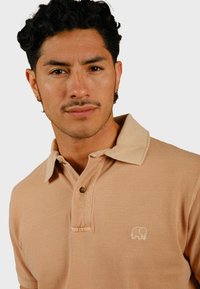 Lichtbeige poloshirt gemaakt van textuurstof, met een knoopsluiting bij de kraag en een klein wit olifantenlogo op de borst.