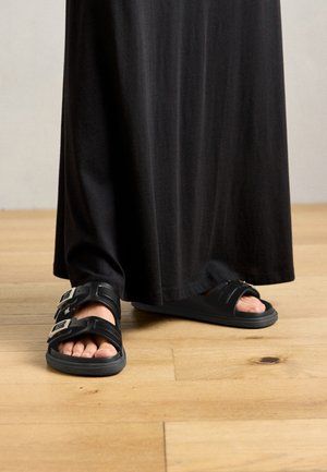 Black sandals with double buckle straps, flat rubber sole, and open toe. Soft texture, minimalistic design, paired with a long black garment.