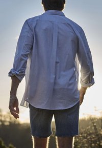 Hombre con camisa de rayas de manga larga y pantalones cortos de mezclilla, de pie al aire libre con luz solar detrás de él.