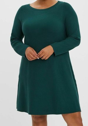 Femme portant une robe verte foncée à manches longues, longueur genoux, avec un col rond, les mains jointes devant sur un fond uni.