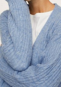 Cardigan en maille torsadée bleu clair avec manches longues, fabriqué en mélange de laine douce et texturée, présentant un col en V profond et une coupe décontractée.