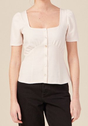 Femme portant un haut blanc à manches courtes boutonné avec un décolleté carré et un pantalon noir taille haute sur un fond uni.