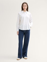 Weißes Hemd mit Knopfleiste im Polka-Dot-Muster, kombiniert mit locker sitzenden, dunkelblauen Jeans und hellen Schuhen vor einem neutralen Hintergrund.