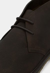 Chaussure en cuir marron présentant une texture lisse, un bout arrondi et deux œillets avec des lacets assortis. Semelle naturelle avec des détails cousus.