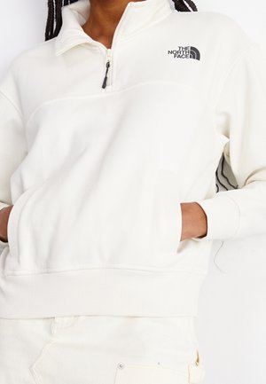 Personne portant un sweat-shirt blanc à quart de fermeture éclair avec une poche frontale et le logo The North Face sur la poitrine, les mains dans la poche.