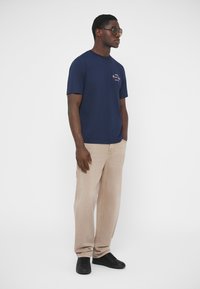 T-shirt en coton bleu marine à col rond, avec une broderie latérale ; associé à un jean beige clair et des baskets noires.