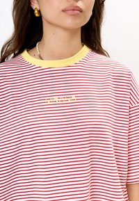 Rood-wit gestreept T-shirt met een gele geribbelde halslijn en gele logotekst. Korte mouwen en een relaxte pasvorm. Zachte katoenmateriaal.
