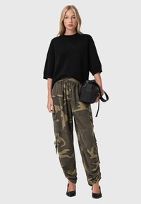 Donna che indossa una maglietta nera oversize a maniche corte, pantaloni cargo mimetici, tacchi neri, mentre tiene una borsa nera su uno sfondo grigio.